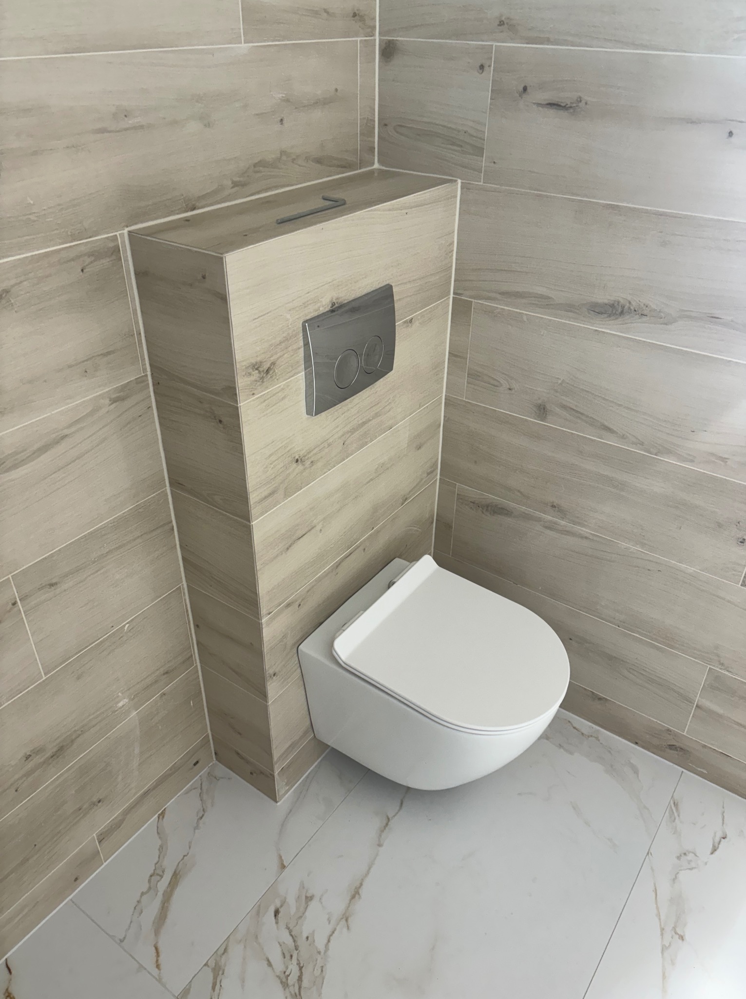Wisząca toaleta z zabudowaną spłuczką, wykończona płytkami imitującymi drewno, na podłodze białe płytki z marmurowym wzorem. Nowoczesna aranżacja łazienki.