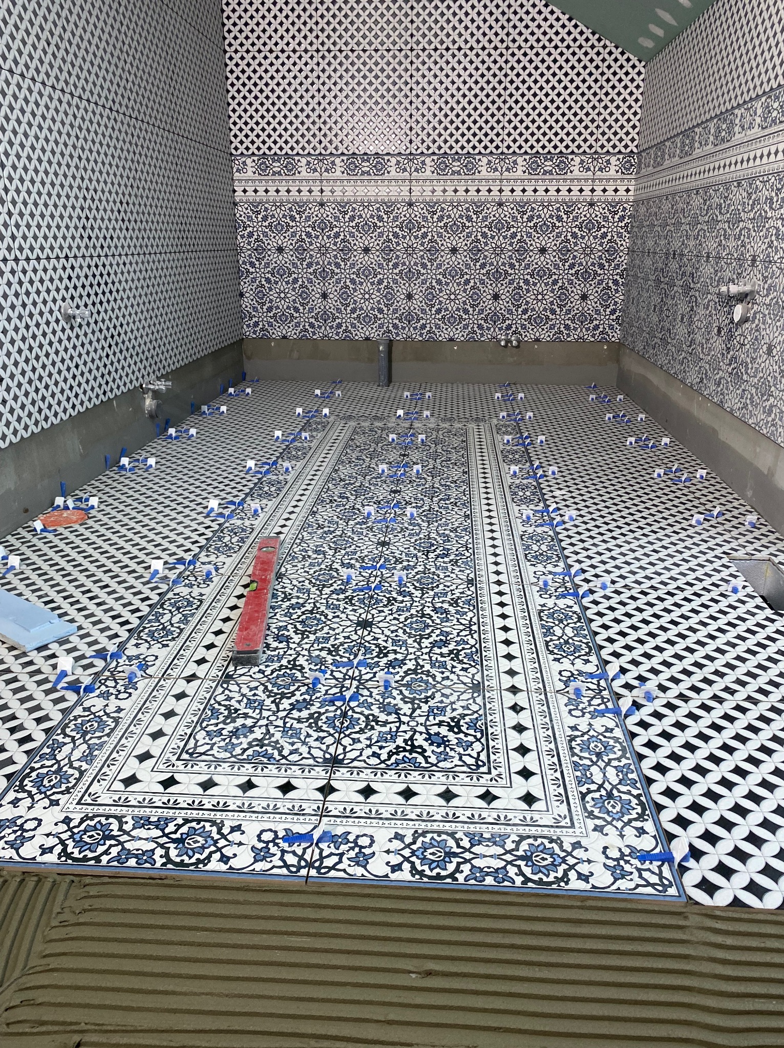 Układanie wzorzystych płytek w łazience, widoczne poziomica i klipsy dystansowe. Na ścianach również płytki z motywami. Etap prac wykończeniowych.