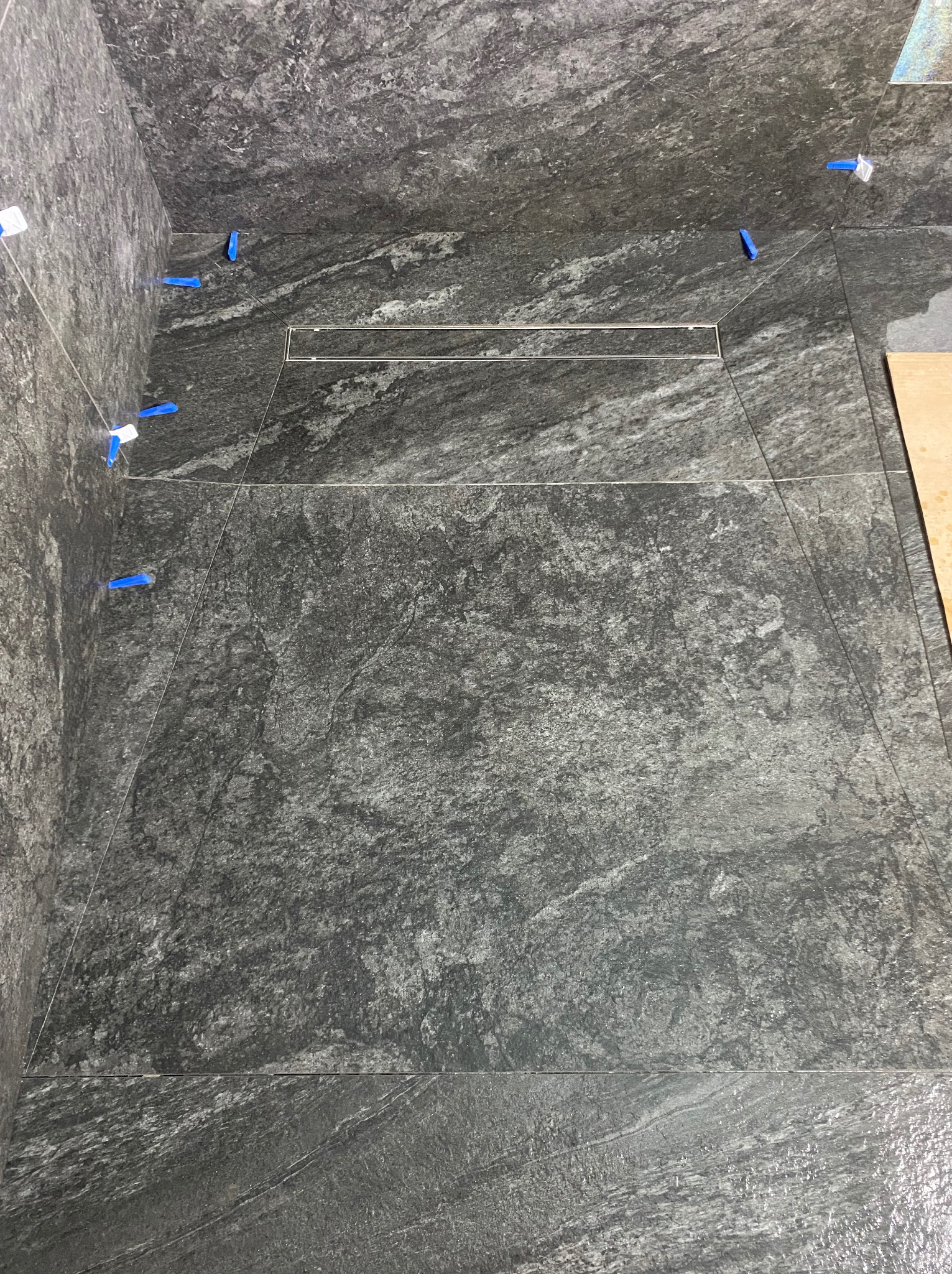 Ułożone, szare płytki imitujące kamień w kabinie prysznicowej z odpływem liniowym. Widoczne niebieskie krzyżyki dystansowe. Częściowo widoczna płyta z boku.