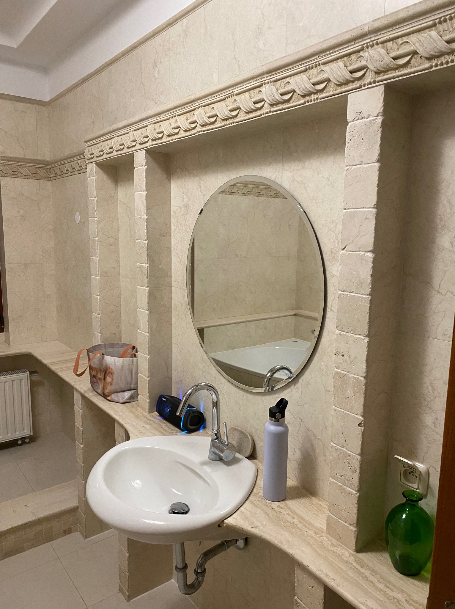 Beżowa łazienka z umywalką, okrągłym lustrem i dekoracyjnymi niszami. Widoczna bateria, butelka na wodę i głośnik. W lustrze odbija się wanna. Całość utrzymana w jasnej tonacji.