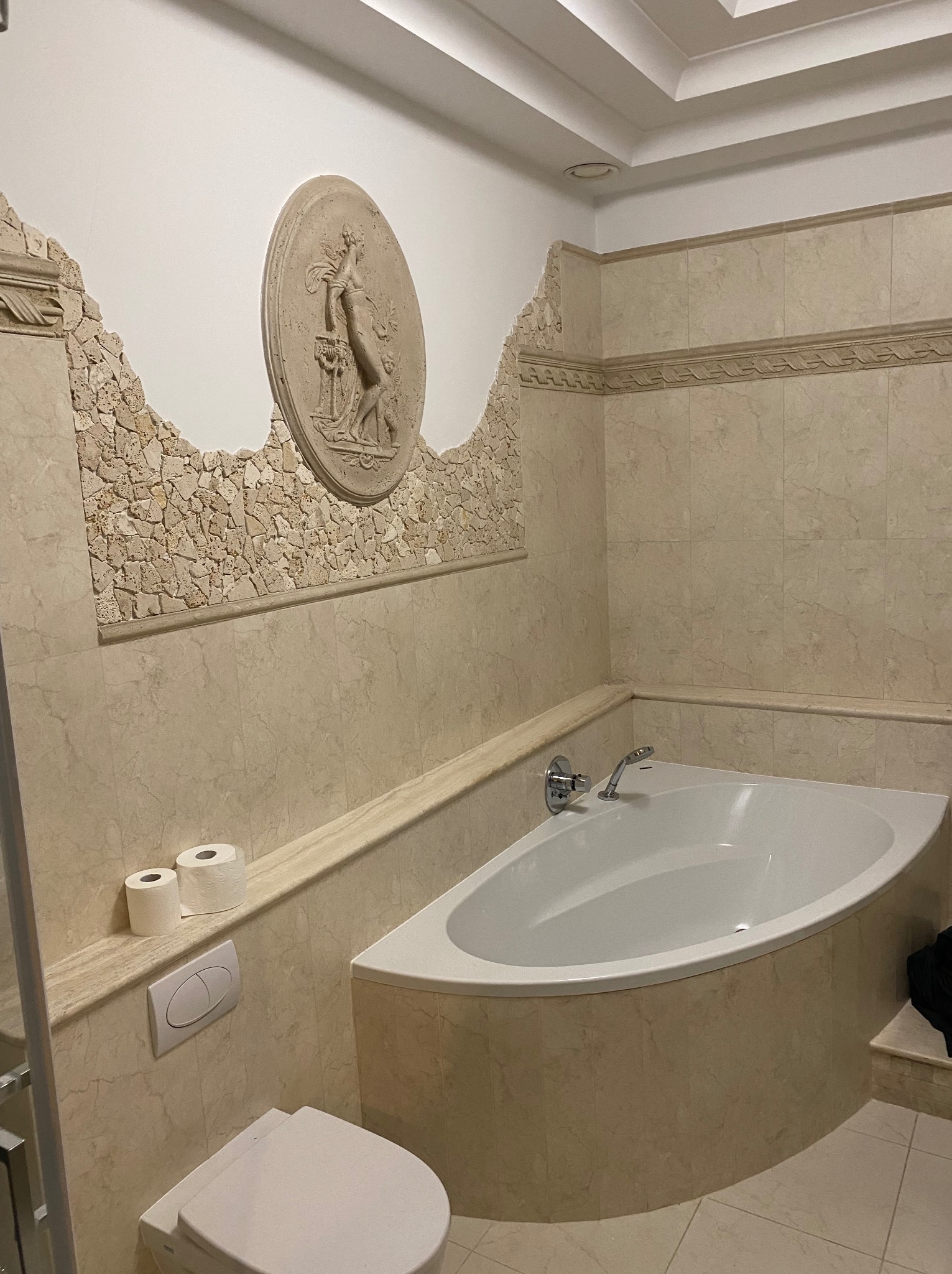 Elegancka łazienka z narożną wanną, beżowymi płytkami i dekoracyjnym panelem ściennym z motywem rzeźby. Widoczna toaleta i fragment kabiny prysznicowej.