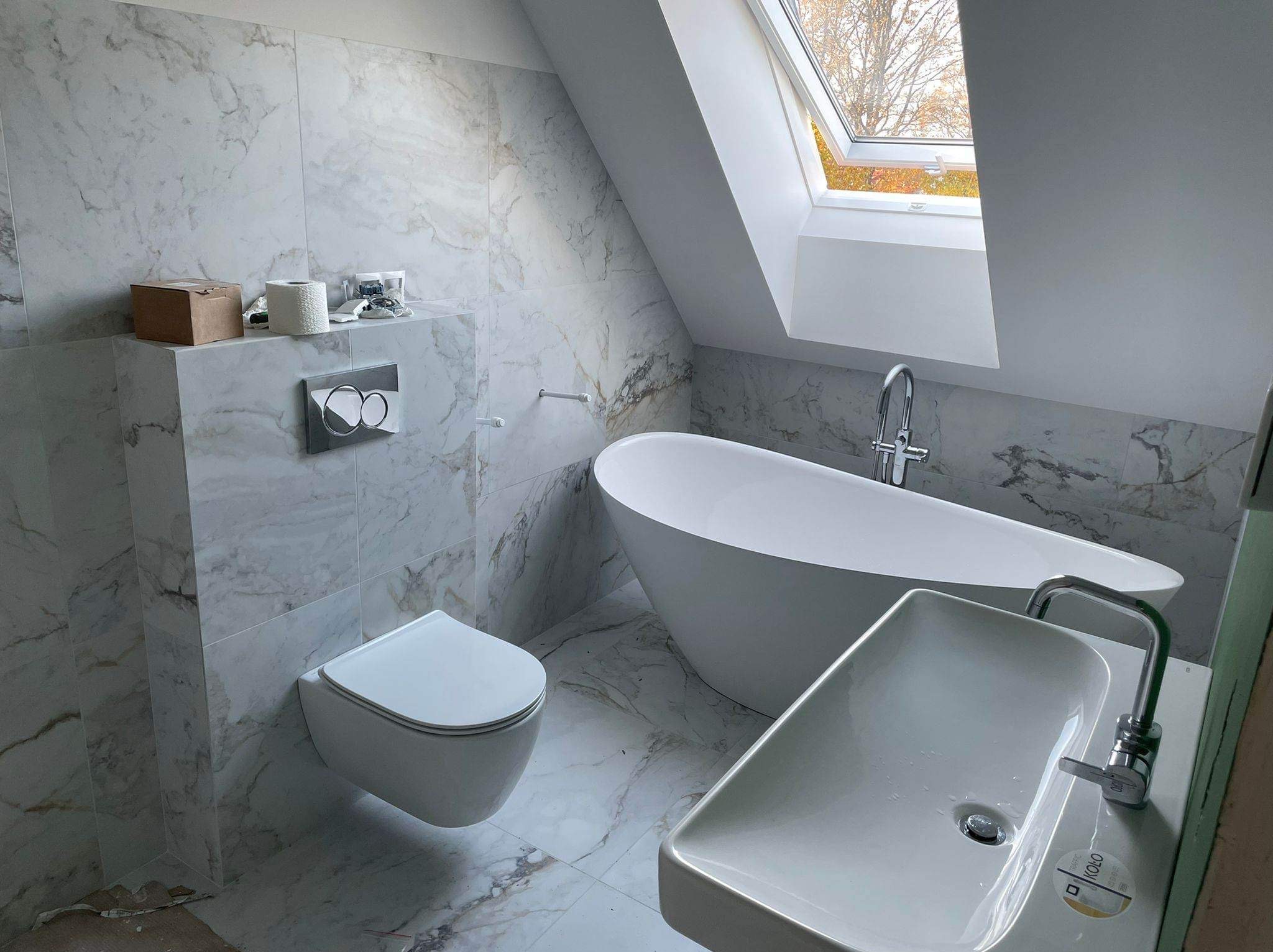 Wykończona łazienka na poddaszu z marmurowymi płytkami, wolnostojącą wanną, podwieszaną toaletą i umywalką KOLO. Okno dachowe wpuszcza naturalne światło.