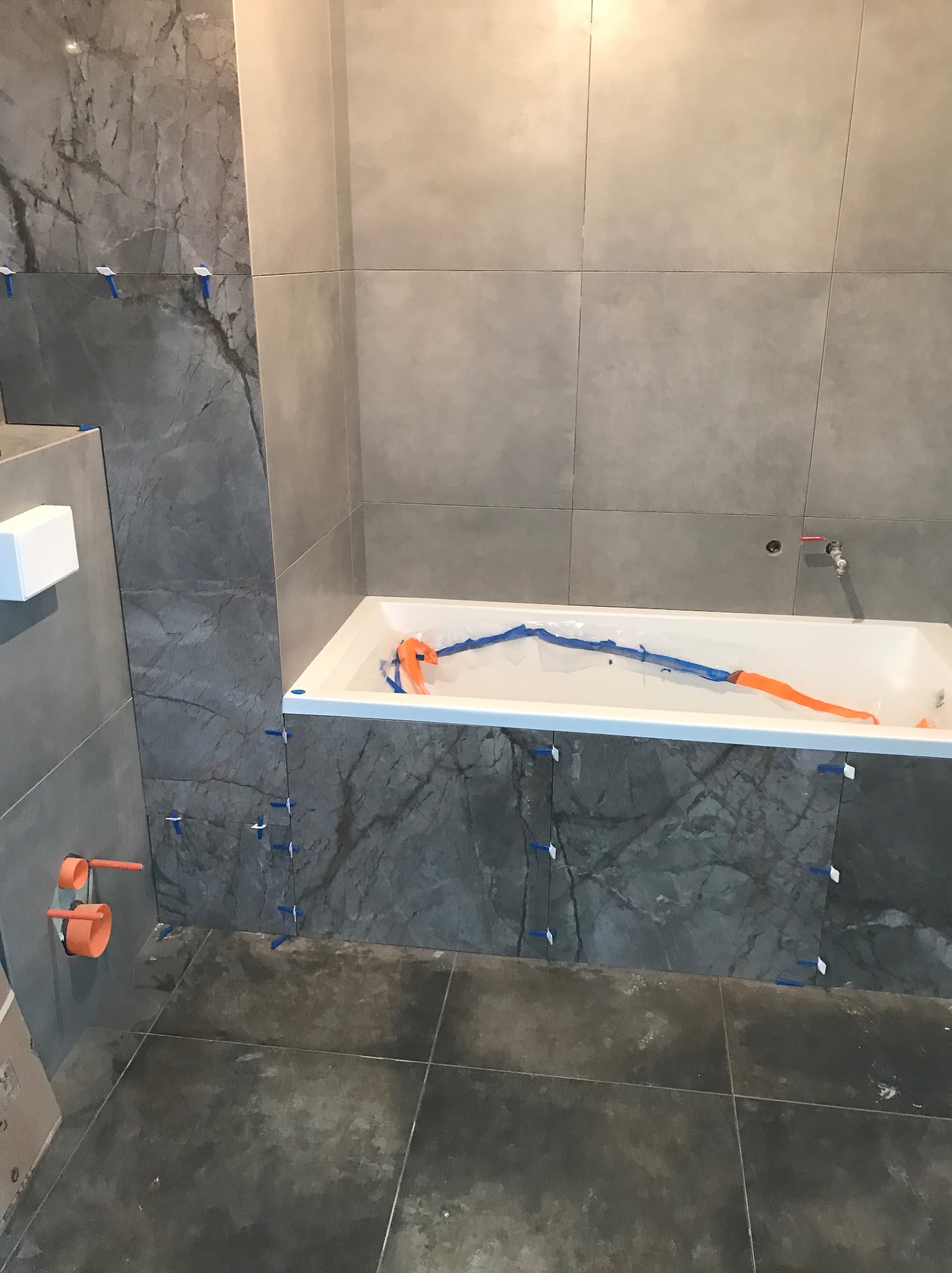 Szare płytki imitujące marmur na ścianach łazienki w trakcie montażu, z krzyżykami dystansowymi. Wanna zabezpieczona folią. Widoczne fragmenty instalacji wodnej.
