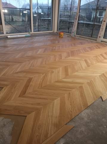 Układanie parkietu w jodełkę w jasnym pomieszczeniu z dużymi oknami. Widoczny fragment surowej podłogi i puszka z klejem. Parkiet z wyraźnym usłojeniem.