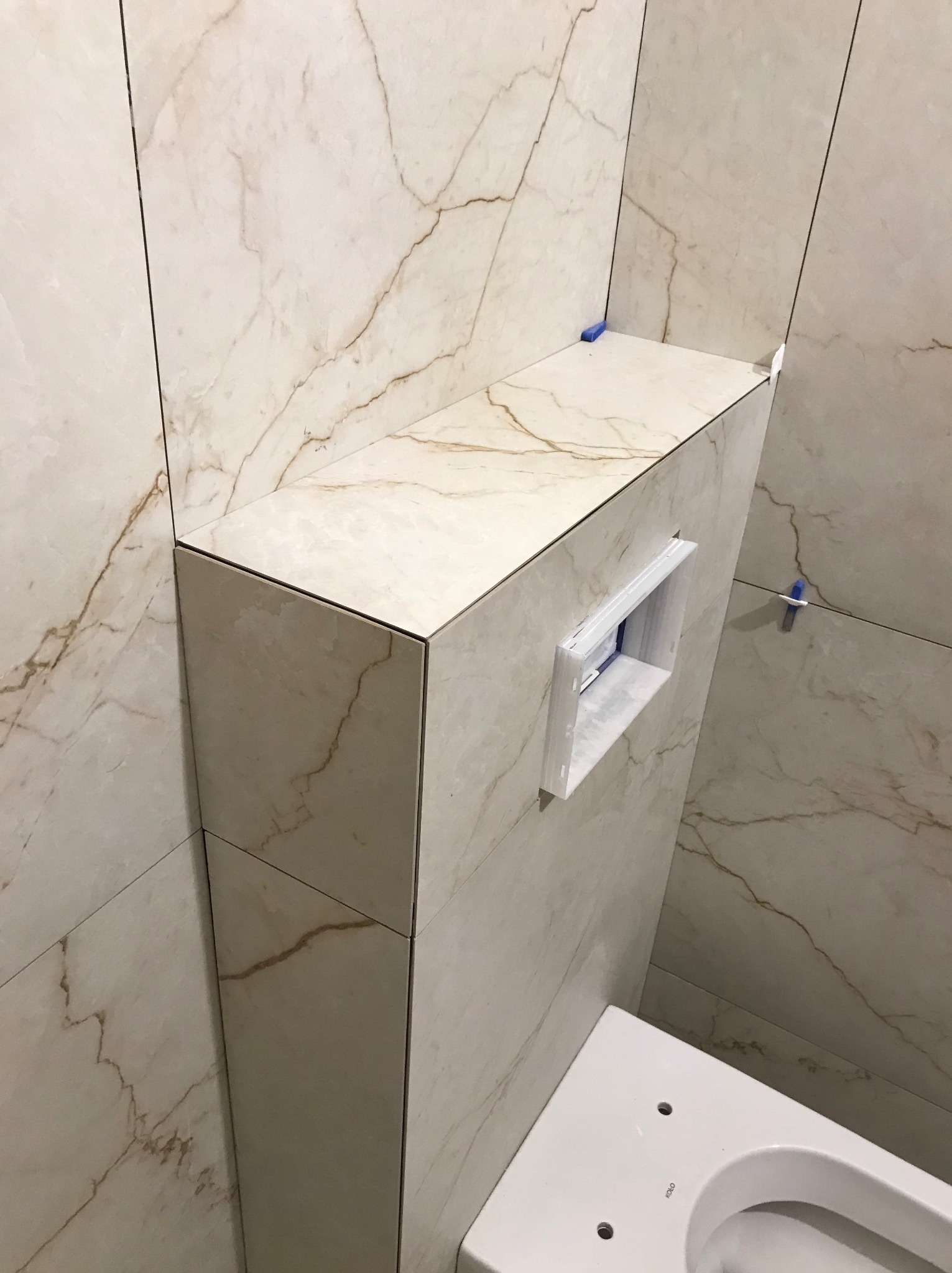 Obudowa stelaża WC z płytek imitujących marmur, z widocznym otworem na przycisk spłukujący. Użyto listew wykończeniowych. Częściowo widoczna miska WC.