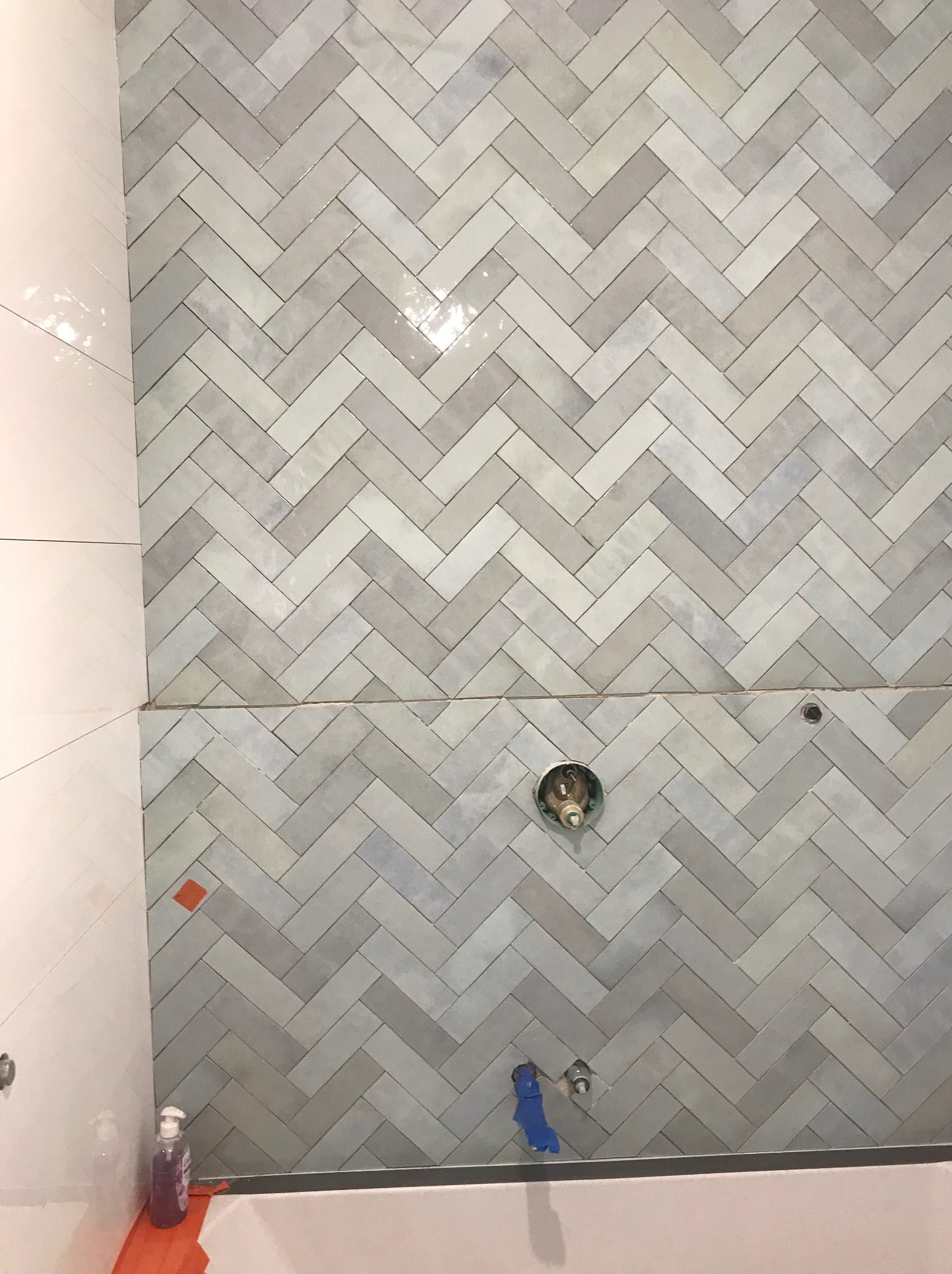 Ściana łazienki z szarymi, ułożonymi w jodełkę płytkami. Widoczne otwory na armaturę i fragment wanny. Częściowo wykończona ściana z białych płytek po lewej.