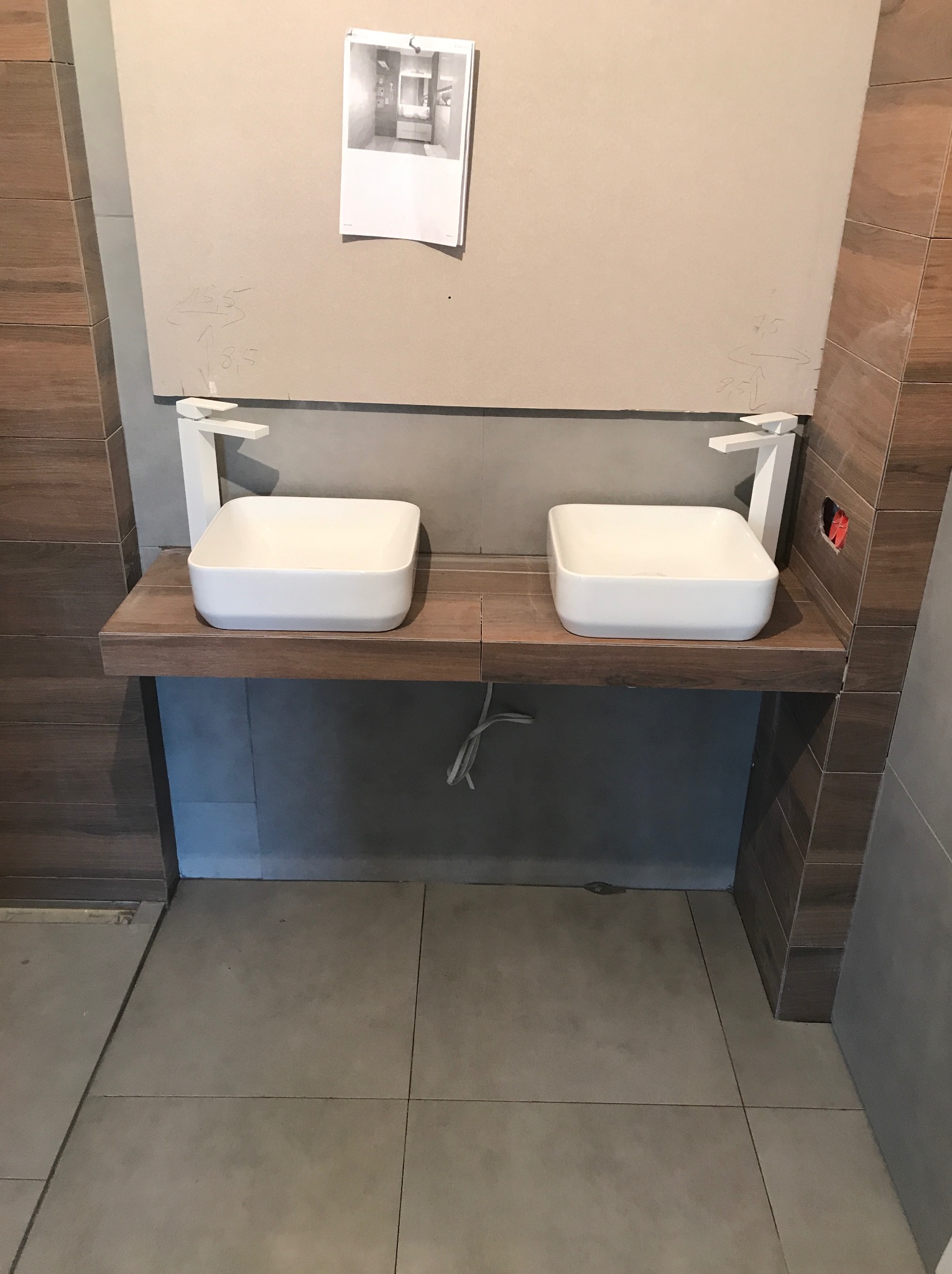 Dwie białe, kwadratowe umywalki nablatowe z nowoczesnymi bateriami na drewnianym blacie w łazience w trakcie wykończenia. Widoczne płytki imitujące drewno i notatki na ścianie.