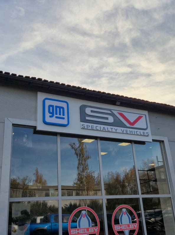 Fasada budynku z dużymi oknami, nad którymi widnieją logotypy 'gm' i 'SV Specialty Vehicles'. W oknach odbija się niebo i drzewa.