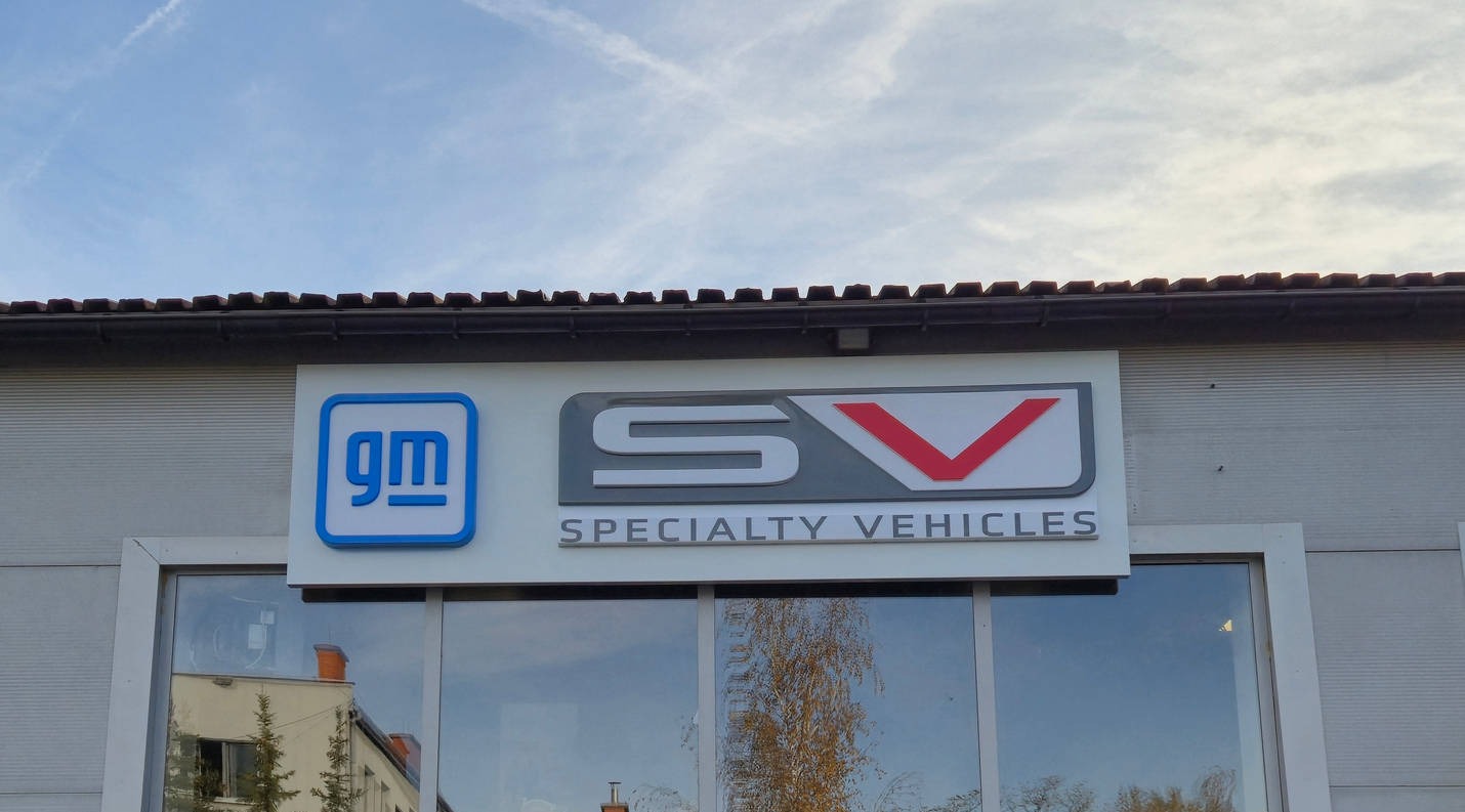 Elewacja budynku z logo GM i SV Specialty Vehicles. Widoczne okna z odbiciem drzewa. Tło: błękitne niebo z chmurami. Ujęcie z dołu, podkreślające wysokość budynku.