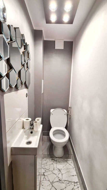 Nowoczesna, wąska łazienka po remoncie: biała toaleta, umywalka z szafką, lustra w kształcie plastra miodu i marmurowy dywanik. Oświetlenie sufitowe.