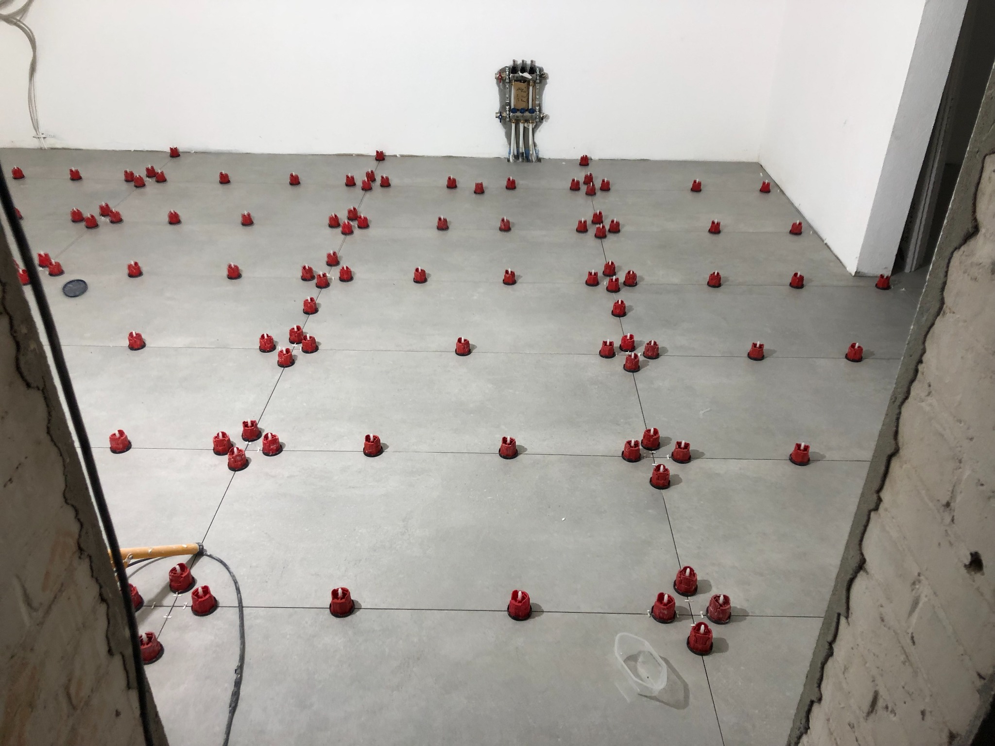 Szare płytki podłogowe z czerwonymi klipsami poziomującymi, ułożone w rzędach z krzyżykami dystansowymi. Widoczne kable elektryczne i fragment ściany z surową cegłą.