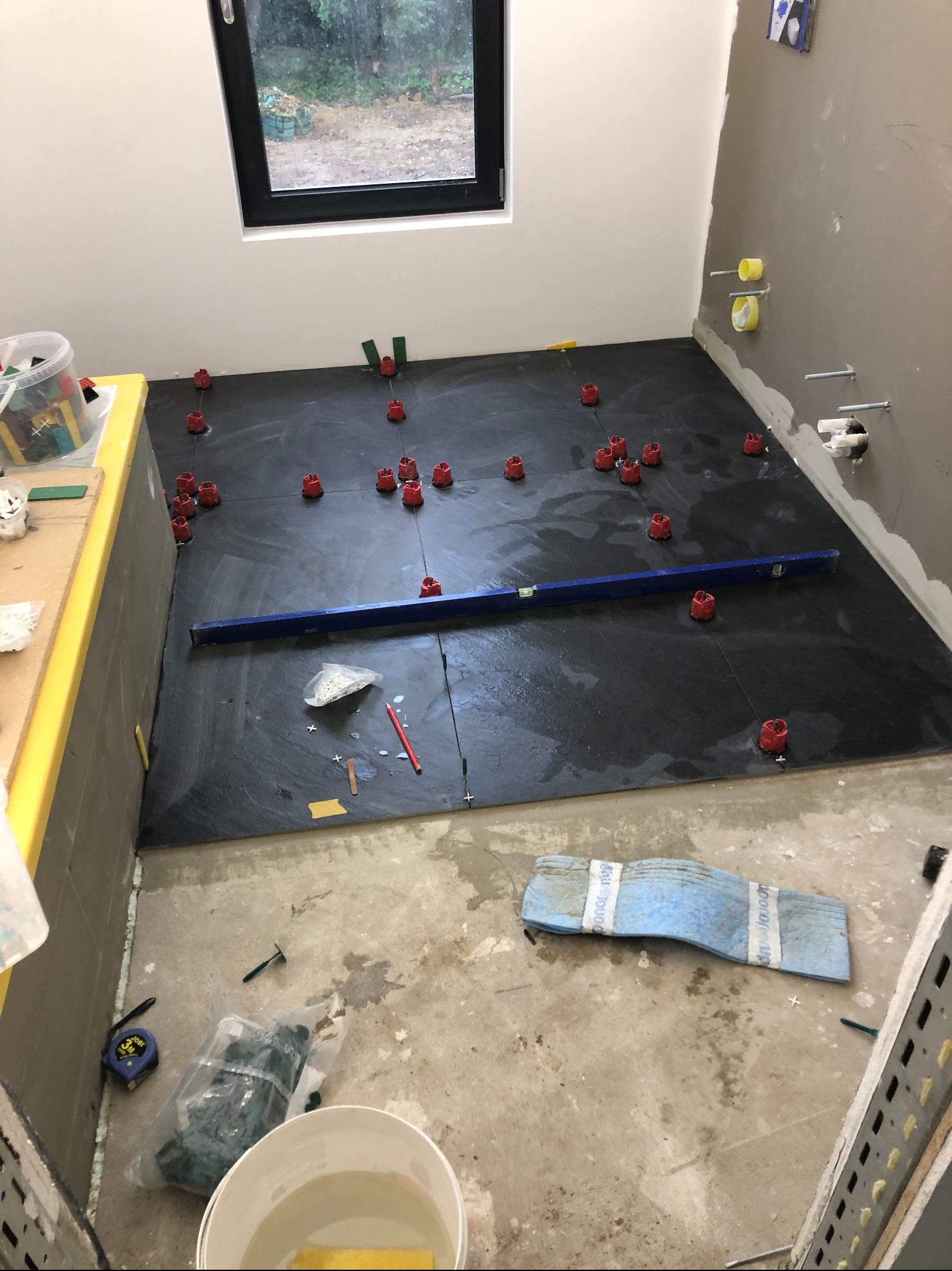 Układanie ciemnych płytek podłogowych w łazience, widoczne poziomica, krzyżyki dystansowe i narzędzia glazurnicze. Okno w tle. Etap prac wykończeniowych.