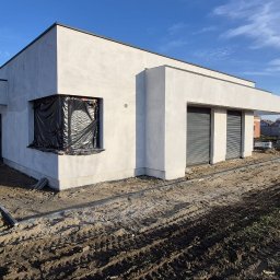 Firma budowlana Matbud - Surowy, otynkowany dom jednorodzinny w trakcie budowy, z zabezpieczonym oknem i przygotowanym terenem wokół. Widoczne ślady opon i materiały budowlane.