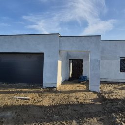 Firma budowlana Matbud - Nowoczesny dom jednorodzinny w trakcie wykończenia elewacji, z widocznym garażem i mini koparką Kubota na posesji. Surowy stan elewacji, błękitne niebo w tle.