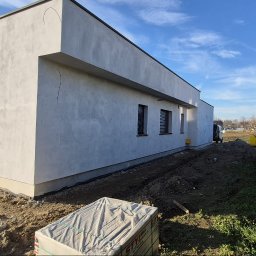 Firma budowlana Matbud - Nowoczesny, parterowy dom z szarą elewacją i płaskim dachem, widoczny z boku. W tle zielony żywopłot i błękitne niebo. Przygotowany teren pod zagospodarowanie.