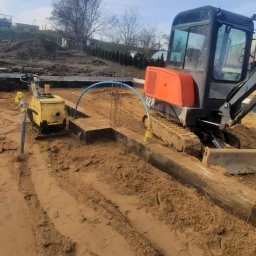 Firma budowlana Matbud - Fundamenty domu w trakcie budowy: wykop, zbrojenie, zagęszczarka i minikoparka na placu budowy. Widoczne niebieskie rury i częściowo ułożone bloczki fundamentowe.