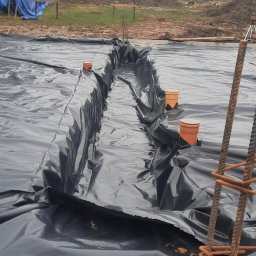 Firma budowlana Matbud - Czarna folia izolacyjna ułożona na fundamencie z widocznymi rurami drenażowymi i zbrojeniem. W tle ziemia i drzewa w pochmurny dzień.
