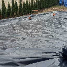 Firma budowlana Matbud - Czarna folia izolacyjna rozłożona na fundamencie domu, widoczne rury wentylacyjne, w tle rząd tuj i budynek z czerwoną dachówką. Etap izolacji fundamentów.