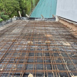 Firma budowlana Matbud - Siatka zbrojeniowa na czarnej hydroizolacji, przygotowana do zalania betonem na dachu budynku. Widoczne cegły i fragment zielonego dachu sąsiedniego budynku.