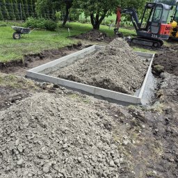 Firma budowlana Matbud - Fundament pod budynek w trakcie budowy: betonowa rama, ziemia w środku, minikoparka Bobcat 328 w tle, taczka na trawie. Widok z góry na plac budowy.
