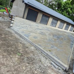 Firma budowlana Matbud - Ułożona kostka brukowa przed budynkiem garażowym z sześcioma bramami, widoczny mini-ładowarka Bobcat i hałda piasku. Krawężniki oddzielają kostkę od gruntu.