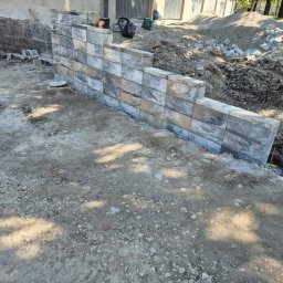 Firma budowlana Matbud - Wzniesiona ściana oporowa z szarych, cieniowanych bloczków betonowych, widoczna w trakcie budowy, z ziemią i gruzem w tle. Ujęcie z dołu, podkreślające strukturę muru.