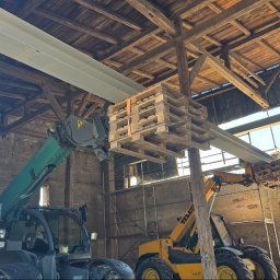 Firma budowlana Matbud - Montaż stalowych belek wewnątrz starego budynku z użyciem dwóch podnośników teleskopowych i palet. Widoczny fragment ściany z cegły i drewniana konstrukcja dachu.