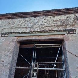 Firma budowlana Matbud - Ceglany budynek w trakcie remontu z widocznym rusztowaniem i stalową belką nad otworem. Częściowo otynkowana ściana i błękitne niebo w tle.