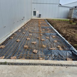 Firma budowlana Matbud - Przygotowanie podłoża: czarna folia, siatka zbrojeniowa i ceglane podkładki na betonowej wylewce przy budynku z blachy trapezowej. Widoczny drenaż liniowy.