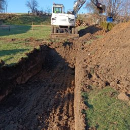 Firma budowlana Matbud - Wykop pod fundamenty wykonany koparką na tle zielonej łąki. Widoczne drewniane znaczniki i sterta drewna. Praca w toku, ziemia świeżo naruszona.