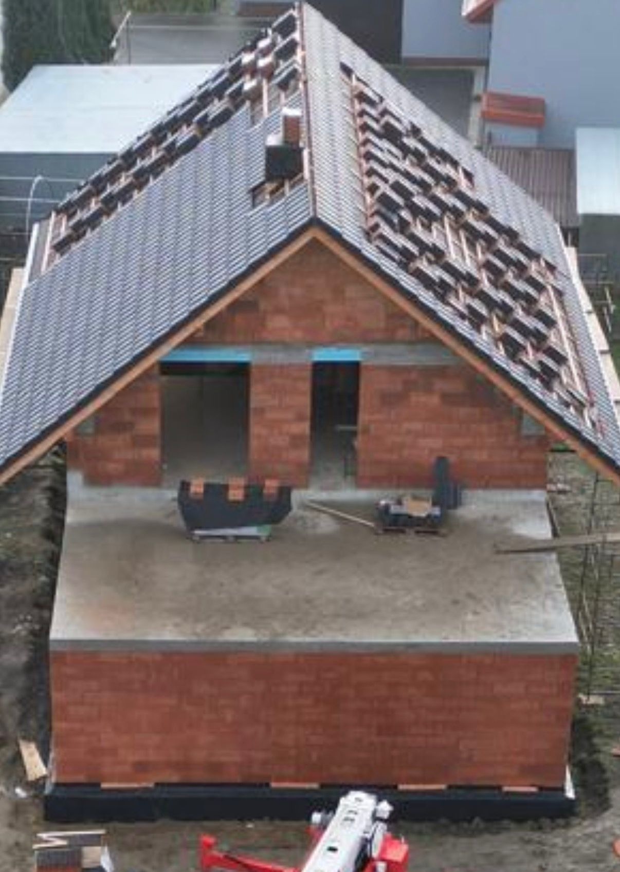 Budowa domu z czerwonej cegły: częściowo pokryty dach z szarej dachówki, widoczne otwory okienne i drzwiowe, fundamenty. Etap wznoszenia ścian i dachu.