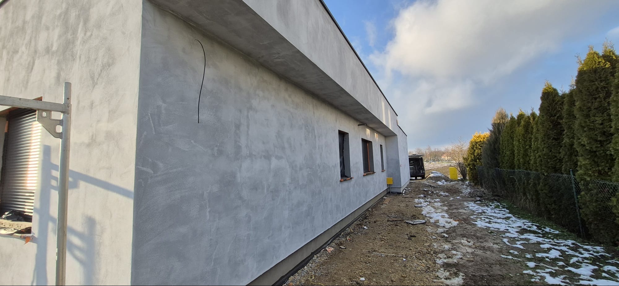 Ściana budynku po nałożeniu szarego tynku strukturalnego, widoczne okna z roletami zewnętrznymi, fragment rusztowania i kabel wystający ze ściany. Częściowo ośnieżone podłoże.