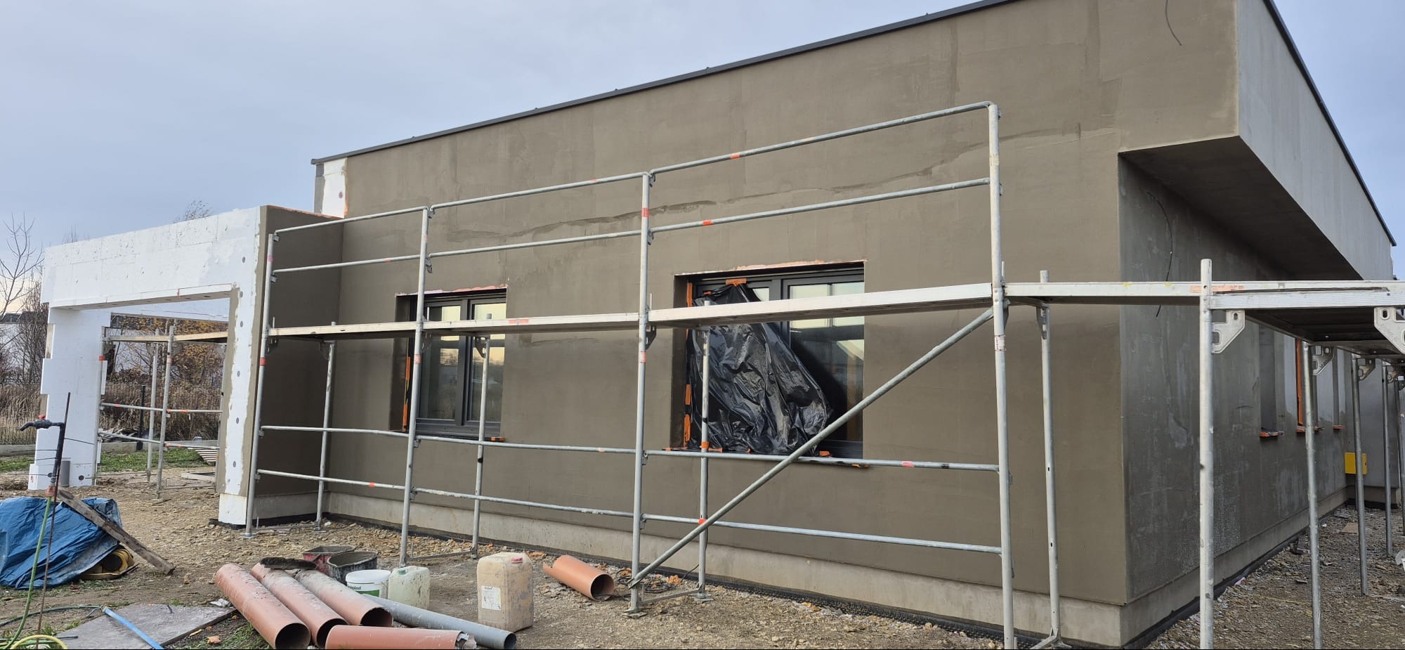 Surowy stan domu jednorodzinnego w Baborowie, z nałożonym tynkiem i rusztowaniem. Okna zabezpieczone folią. Widoczne elementy izolacji i rury kanalizacyjne.