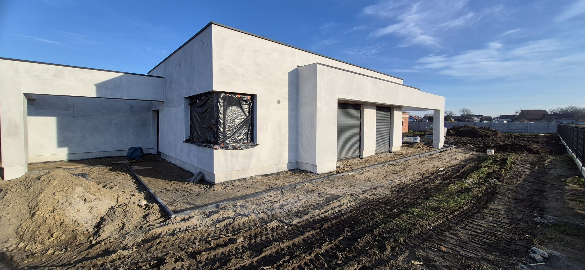 Surowy, otynkowany dom jednorodzinny w trakcie budowy, z zabezpieczonym oknem i przygotowanym terenem wokół. Widoczne ślady opon i materiały budowlane.