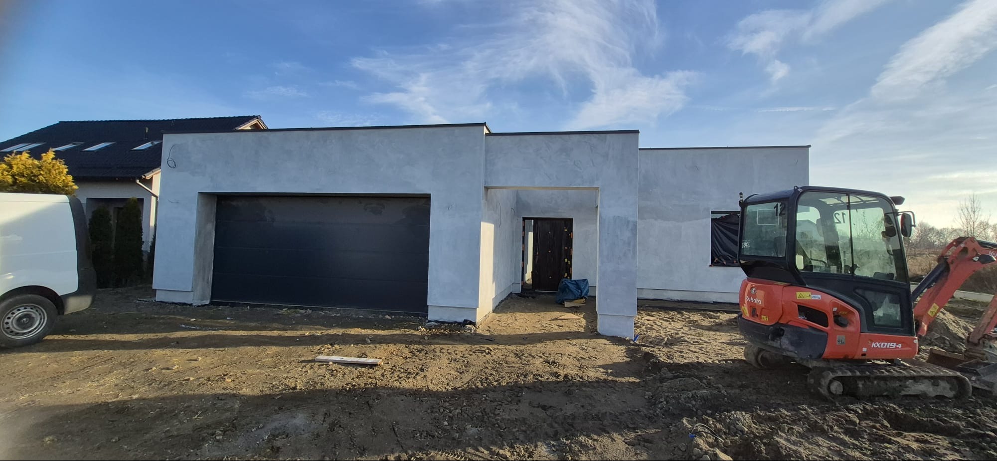 Nowoczesny dom jednorodzinny w trakcie wykończenia elewacji, z widocznym garażem i mini koparką Kubota na posesji. Surowy stan elewacji, błękitne niebo w tle.