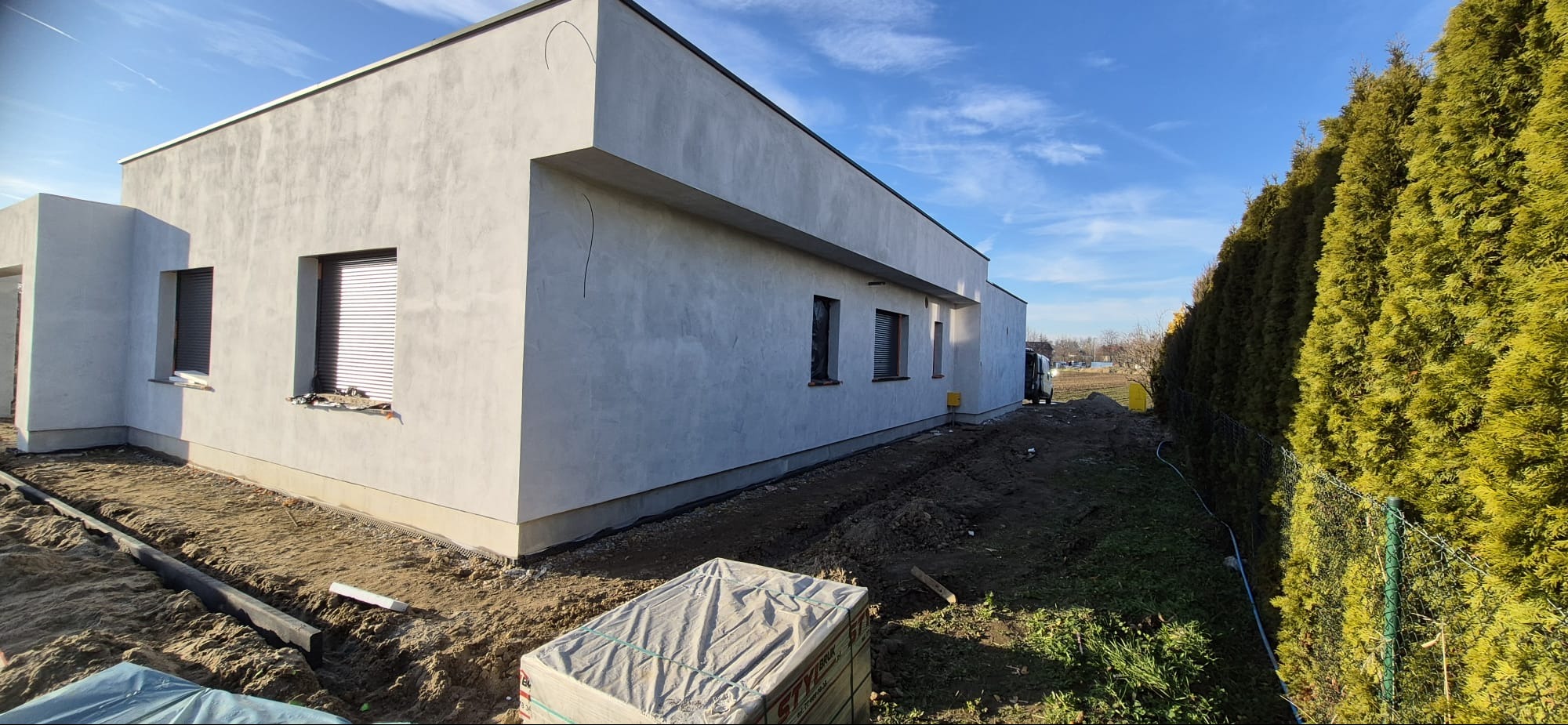 Nowoczesny, parterowy dom z szarą elewacją i płaskim dachem, widoczny z boku. W tle zielony żywopłot i błękitne niebo. Przygotowany teren pod zagospodarowanie.