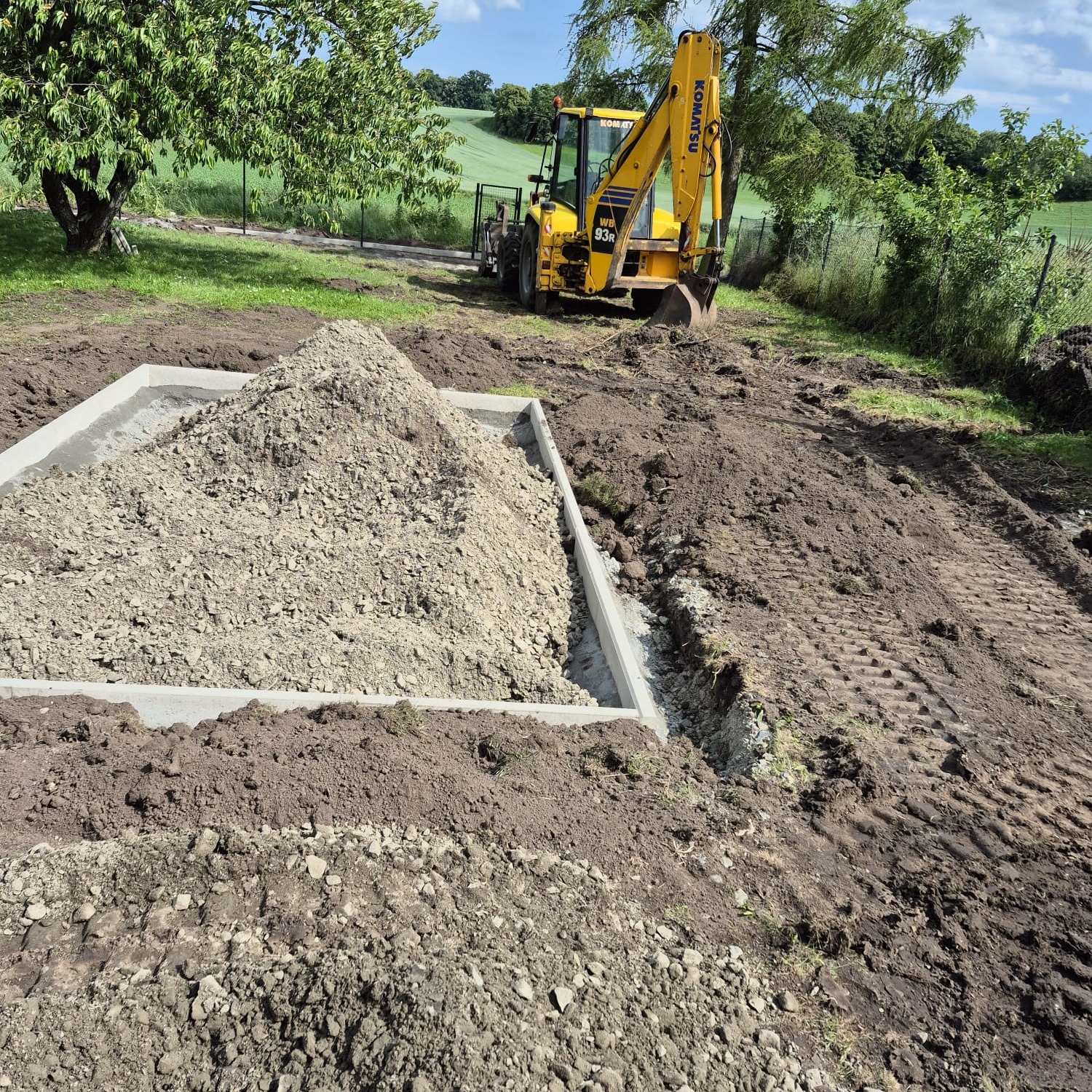 Fundament z betonową wylewką, zasypany żwirem, z koparką Komatsu w tle na zielonym terenie. Widok z góry na plac budowy.