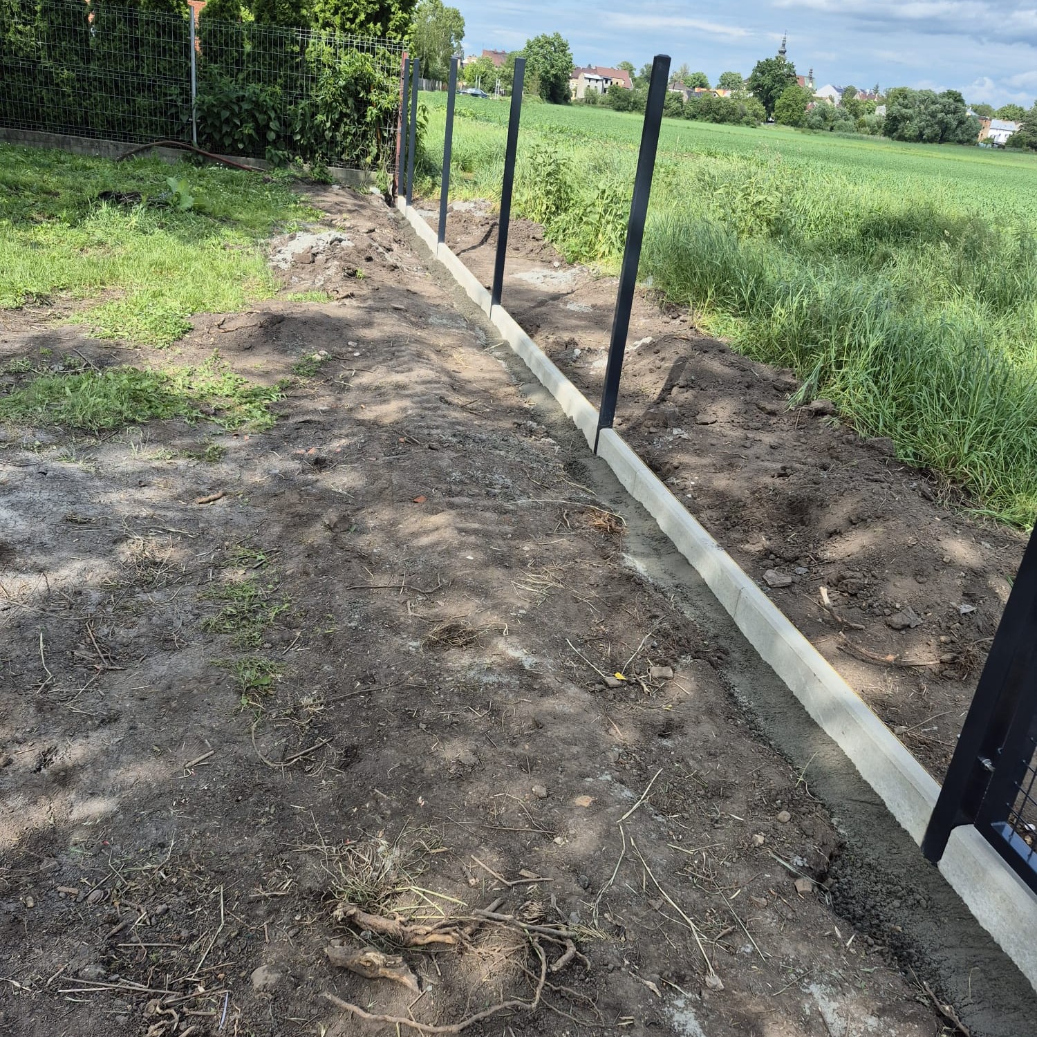 Betonowe obrzeża w gruncie, przygotowane pod montaż ogrodzenia panelowego. Widoczne słupki i zielone tło z zabudowaniami w oddali.