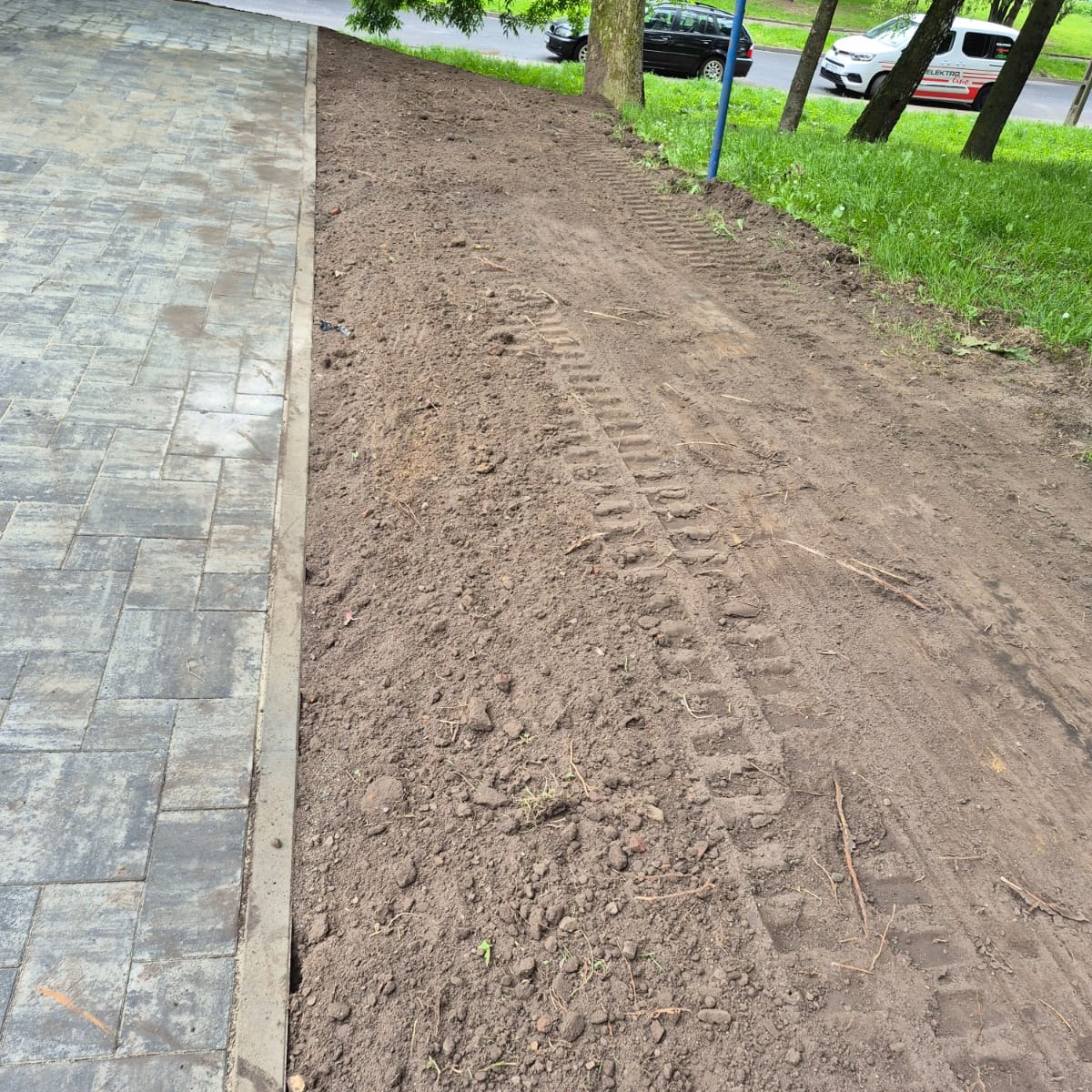 Ułożona kostka brukowa sąsiaduje z przygotowanym podłożem pod trawnik, widoczne ślady opon. Prace ziemne w toku, teren wyrównany.