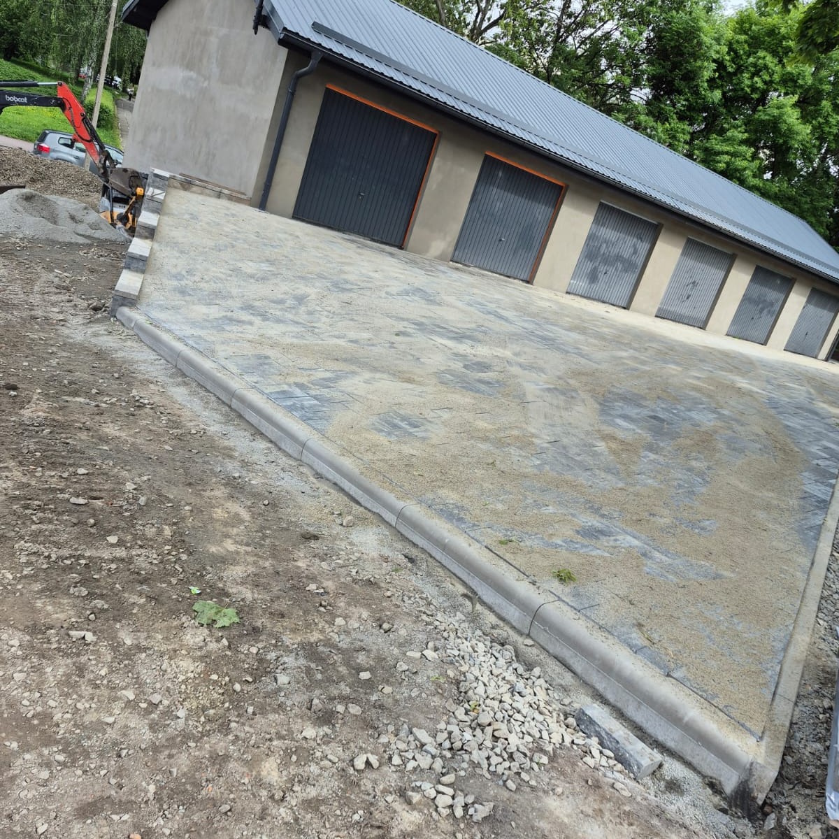 Ułożona kostka brukowa przed budynkiem garażowym z sześcioma bramami, widoczny mini-ładowarka Bobcat i hałda piasku. Krawężniki oddzielają kostkę od gruntu.