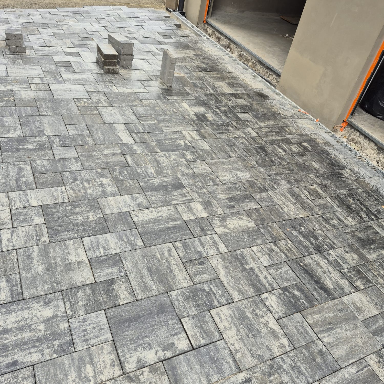 Układanie szarej kostki brukowej na podjeździe, widoczne niedokończone fragmenty i stosy kostki, zarys budynku w tle. Charakterystyczny wzór kostki.