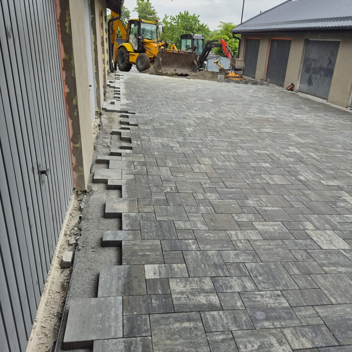 Układanie szarej kostki brukowej na podjeździe, z widocznymi maszynami budowlanymi w tle. Częściowo ułożona nawierzchnia, etap prac.