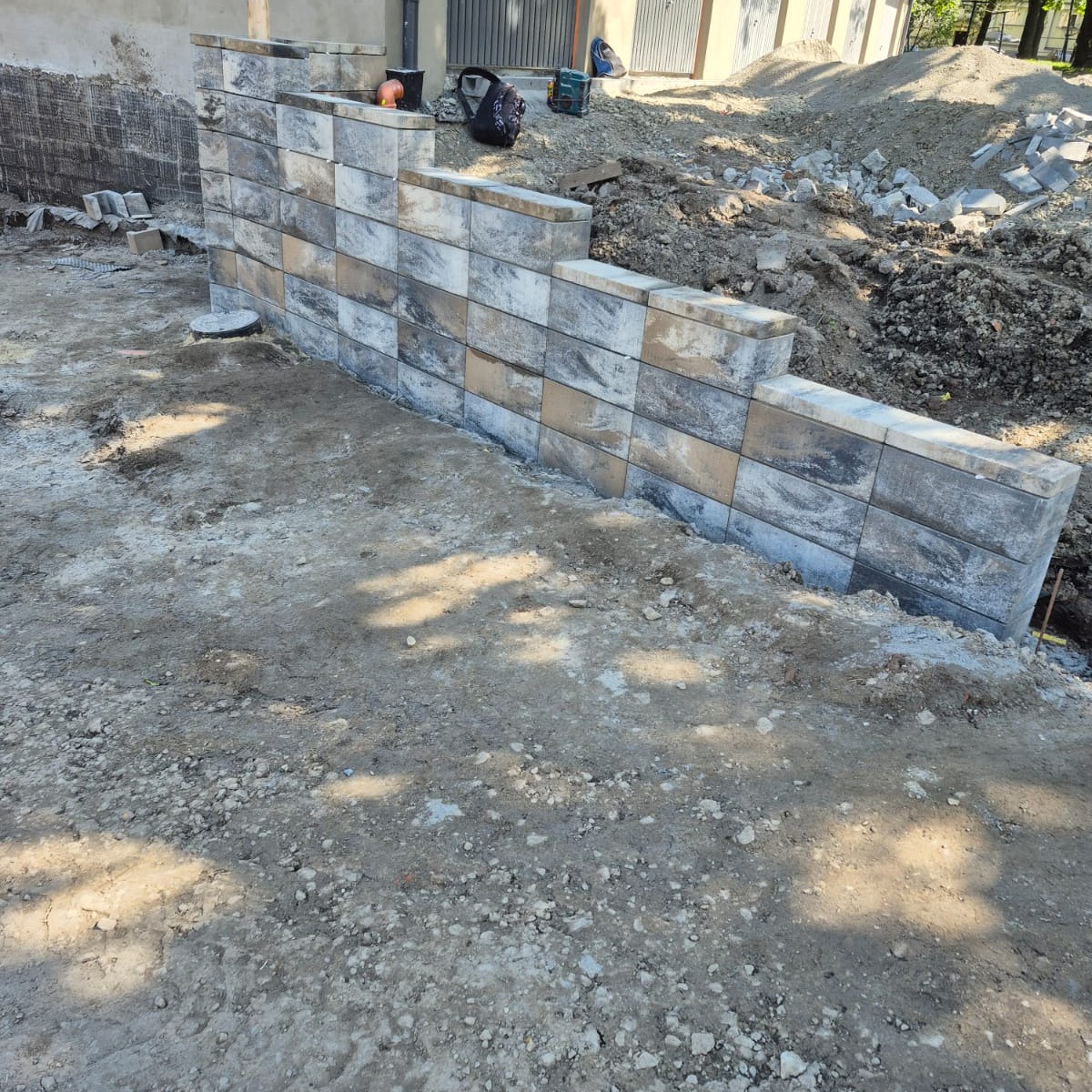 Wzniesiona ściana oporowa z szarych, cieniowanych bloczków betonowych, widoczna w trakcie budowy, z ziemią i gruzem w tle. Ujęcie z dołu, podkreślające strukturę muru.