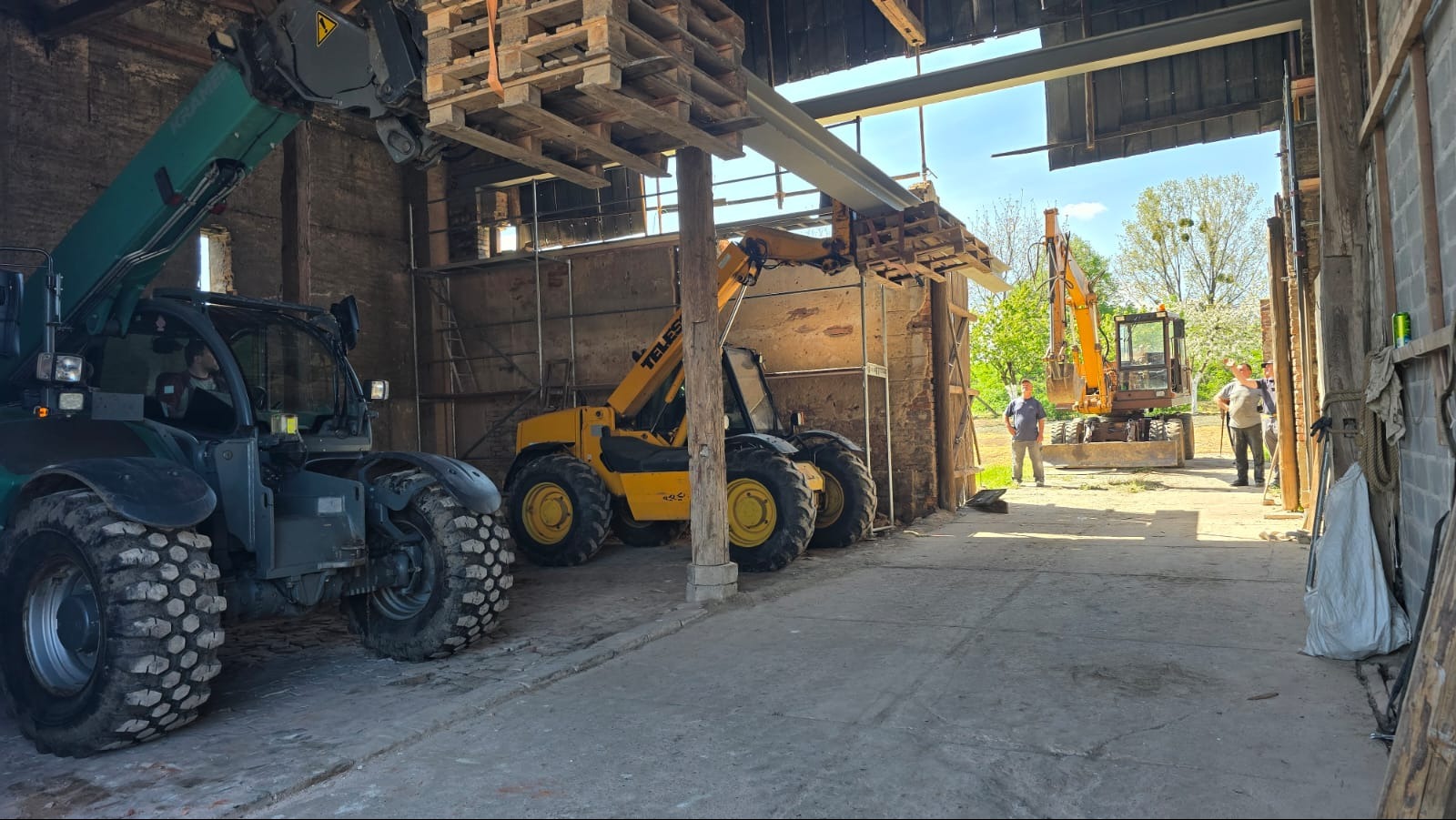 Wnętrze remontowanego budynku z maszynami budowlanymi: podnośniki teleskopowe i koparka. Widoczni pracownicy. Elementy konstrukcyjne w trakcie montażu.