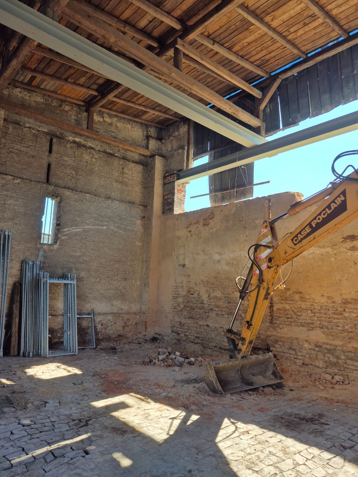 Wnętrze starego budynku w trakcie przebudowy z widocznym sprzętem budowlanym, rusztowaniami i elementami konstrukcyjnymi dachu.