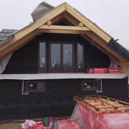DC Okna - Dariusz Chlebek - Budowa domu w trakcie: widoczne okna z brązowymi ramami, drewniana konstrukcja dachu, czarna membrana i ocieplenie Rockwool. Etap prac wykończeniowych.