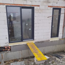 DC Okna - Dariusz Chlebek - Montaż okien przesuwnych i standardowych w surowym budynku z białego bloczka. Widoczne żółte deski szalunkowe i fragmenty ocieplenia fundamentów.