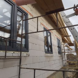 DC Okna - Dariusz Chlebek - Montaż okien PCV w surowym budynku z białego bloczka. Widoczne rusztowanie, drabina i piła łańcuchowa. Okna w ciemnych ramach, uszczelnione pianką montażową.