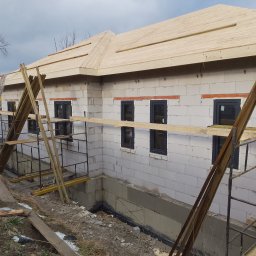 DC Okna - Dariusz Chlebek - Budowa domu z bloczków: wstawione okna z czarnymi ramami, drewniana konstrukcja dachu w trakcie montażu, rusztowania i materiały budowlane wokół.