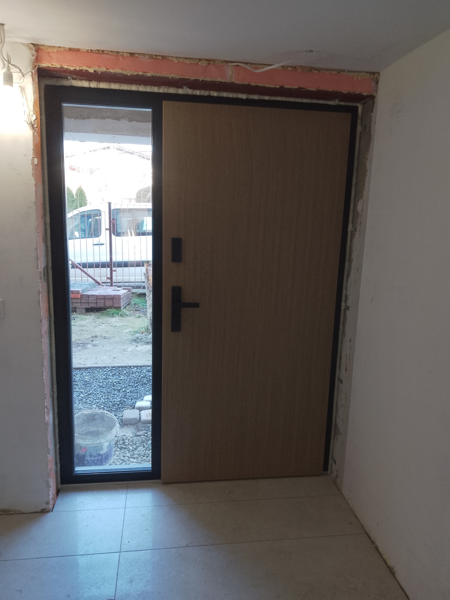 Montaż drzwi zewnętrznych: drewniane drzwi z czarną ramą w trakcie montażu, widoczne materiały budowlane i otoczenie zewnętrzne przez otwartą część ramy.